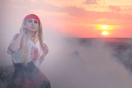 Beautiful young blonde girl with a red scarf, a traditional romanian blouse and a red and black skirt covered in smoke at sunset, Romaniaの写真素材