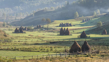 Beautiful rural mountain landscape in the morning light with fog,old houses and haystacks, Fundatura Ponorului, Hunedoara County, Romaniaの写真素材