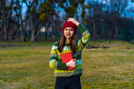 A teenager girl in red hat and green striped sweater holding book with red cover while standing on a lawn in a parkの写真素材