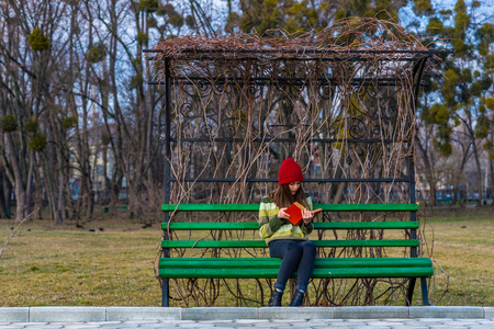 A teenager girl is sitting on a bench in a park and reading book with red cover.の写真素材