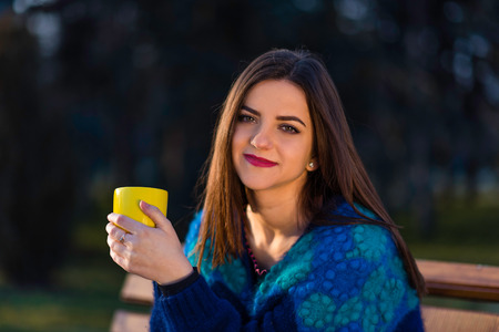 A teenager girl in turquoise blouse is sitting on a bench in a park and holding a yellow cup with two hands.の写真素材