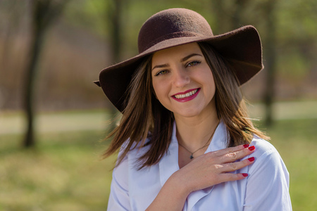 Portrait of a smiling young woman, holding her hand on her shoulder and wearing a hat in a park during spring. Woman bowed her head head to the left side of frame. Medium shot. Shallow depth of field.の写真素材
