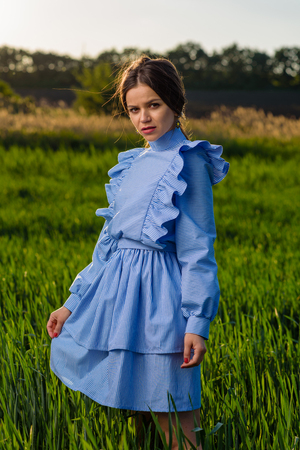 Young woman in blue striped dress is looking towards the camera while standing in the field of green wheat at a spring afternoon during sunny weather. She is holding her dress with one hand. Her hair is fluttering in the wind.の写真素材