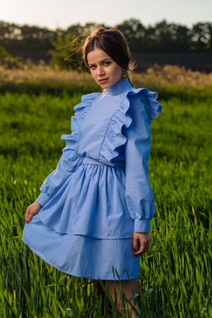 Young slightly smiling woman in blue and white striped dress is looking towards the camera while standing in the field of green wheat at a spring afternoon during sunny weather. She is holding her dress with one hand. Her hair is fluttering in the wind.の写真素材