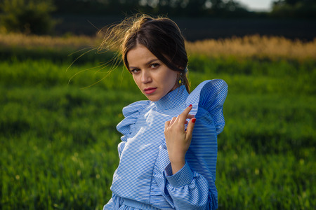 Young woman in blue and white striped dress is looking towards the camera while standing in the field of green wheat at a spring afternoon during sunny weather. She is holding one hand at her shoulder. Her hair is fluttering in the wind.の写真素材