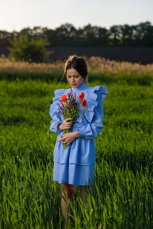 Young woman in blue and white striped dress is looking down, holding a bouquet of summer flowers while standing in the field of green wheat at a spring afternoon during sunny weather. Her hair is fluttering in the wind.の写真素材