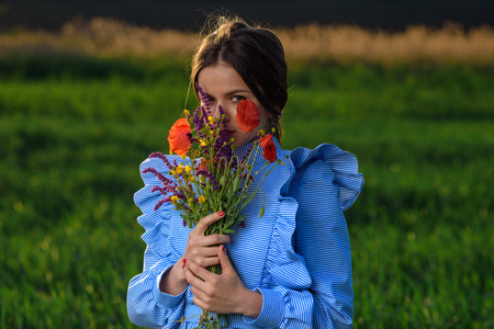 Young woman in blue and white striped dress is holding a bouquet of summer flowers and looking through it to the camera while standing in the field of green wheat at a spring afternoon during sunny weather. Her hair is fluttering in the wind. Girl looks pensive.の写真素材