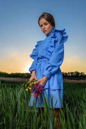 Young woman in blue and white striped dress is holding a bouquet of summer flowers with two hands while standing in the field of green wheat at a spring afternoon during sunny weather and looking to the camera. The sun is setting behind her.の写真素材