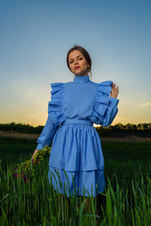 Young woman in blue and white striped dress is holding a bouquet of summer flowers with one hand while standing in the field of green wheat at a spring afternoon during sunset. Girl is looking to the camera, one of her hand is raised to the level of shoulder.の写真素材