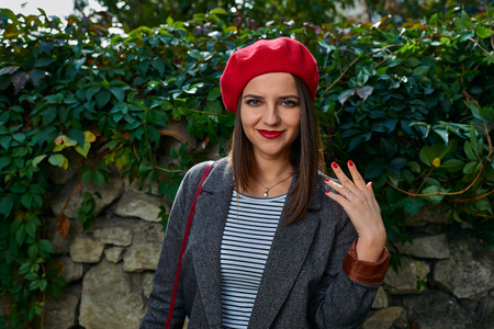 Young woman in red beret is showing red nails near a stone fence covered in vegetation.の写真素材
