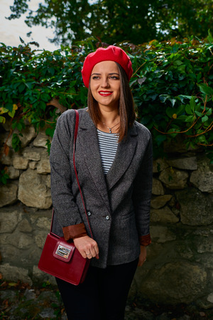 Portrait of a young smiling woman in red beret with red handbag looking away from camera near stone fence covered with vegetation.の写真素材