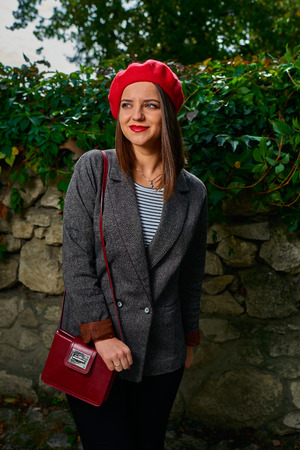 Portrait of a young woman in red beret with red handbag looking away from camera near stone fence covered with vegetation.の写真素材