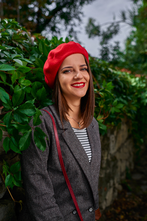 Portrait of a young smiling woman in red beret leaning on a stone fence covered with vegetation.の写真素材
