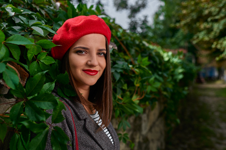 Close up shot of smiling young woman in red beret leaning on a stone fence covered with vegetation.の写真素材