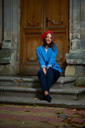 Young woman in red beret and blue jacket is sitting on a concrete porch near wooden doors.の写真素材
