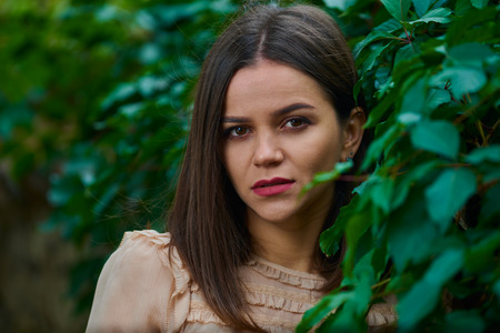 Close up portrait of a young woman leaning on a stone fence covered with green leaves and looking into camera.の写真素材