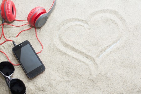 phone with headphones and sunglasses on sand in a summer beach, heart pattern on sandの写真素材