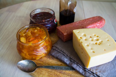 Big piece of cheese, salami and jars with jam. Wooden cutting board.の写真素材