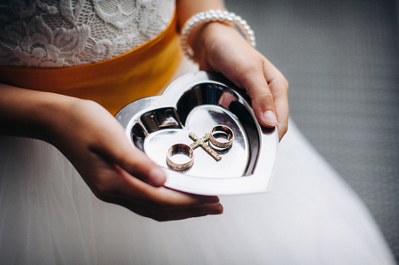 The bride is holding wedding rings on a plate for rings. Bride in a white dress with a yellow belt.の写真素材