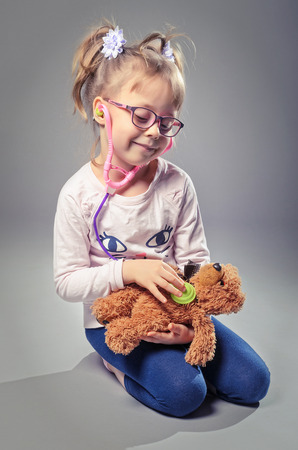 Pretty girl plays in the doctor treats a teddy bear on a gray background. Beautiful girl listens to a stethoscope bear.の写真素材