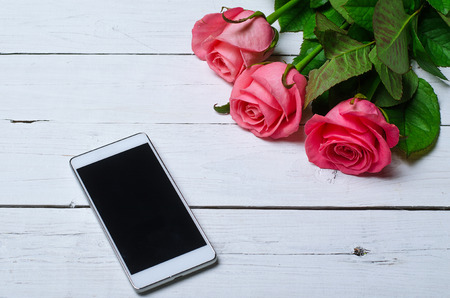 Pink roses and smartphone on a wooden white background. Background for the holiday: valentine's day, birthday, mother's day.の写真素材
