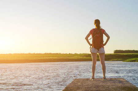 A happy woman is standing on the pier and enjoying the sunset, free space. Young woman with a backpack on the river bank, beautiful landscape.の写真素材