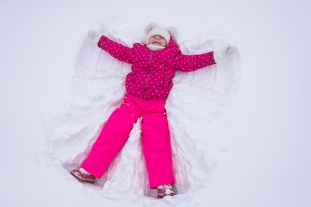 Beautiful little girl makes a snow angel in a park, top view. The child lies in the snow, the winter weather.の写真素材