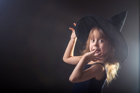 Beautiful girl in a witch hat on a dark background with free space for your text. Portrait of little girl in carnival costume of sorceress, background on halloween.の写真素材