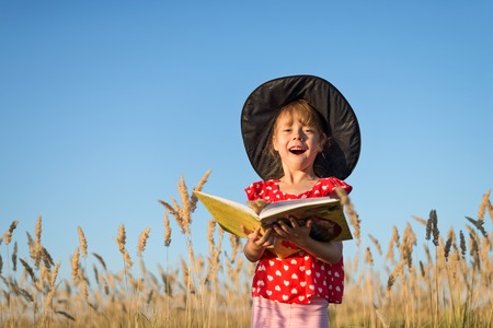 ?hild in a witch hat outdoors reading a book, postcard for the autumn holiday Halloween. Little girl in a witch costume, fabulous background.の写真素材