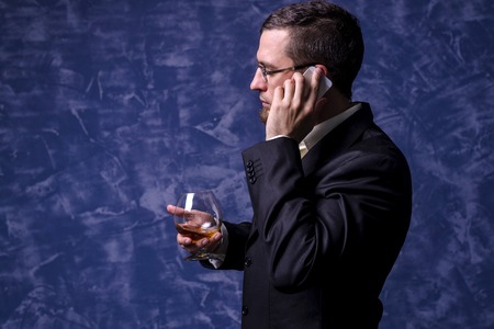 Business man with a glass of brandy and a smartphone. A man in a suit on a blue background.の写真素材