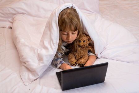 Cute little girl using a laptop in bed under a blanket. A girl is watching a cartoon on a laptop.