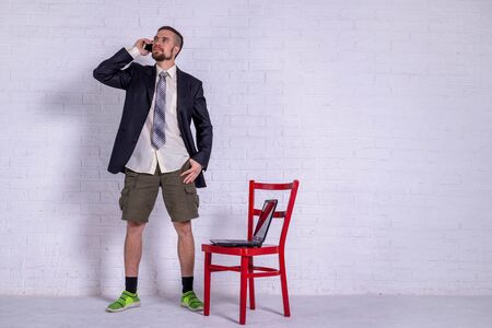 A young man in a suit and shorts is talking on the phone, and a laptop is on the chair. The concept of remote work. Freelanceの写真素材