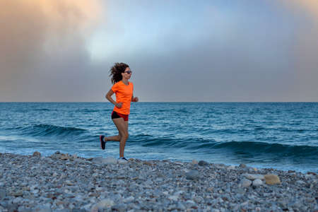 A slender woman in a bright orange T-shirt runs along beach during bad weather.