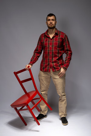 Athletic young man in a red shirt and pants posing with a red chair on a gray backgroundの写真素材