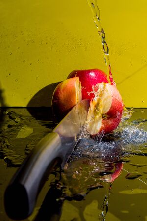 red apples with drops, splashes, slices of apples, water, knife on a yellow background