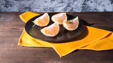 Tangerine slices on a black plate with a yellow napkin on an old natural wooden table, side view,...