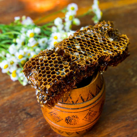 Honeycomb with honey on a clay pot with camomiles on a wooden table, close-up, background, texture,...