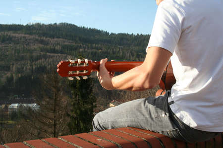 Young man is sitting outside house (on the roof) and is playing.  Mountain scenery