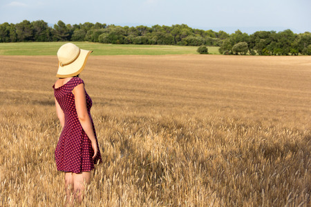 Beautiful girl in sundress and straw hat standing in ripening wheat filed starring in distanceの写真素材