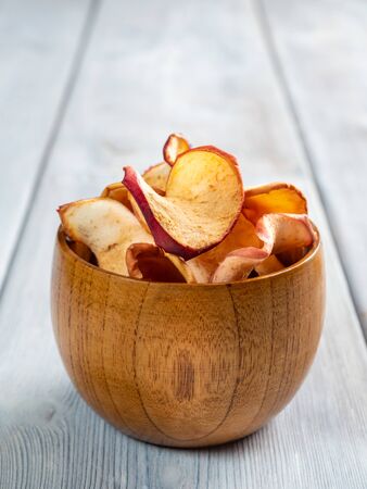 Close up view of tasty apple chips in a wooden cup on the white wooden background. Vertical orientation. Soft focus.の写真素材