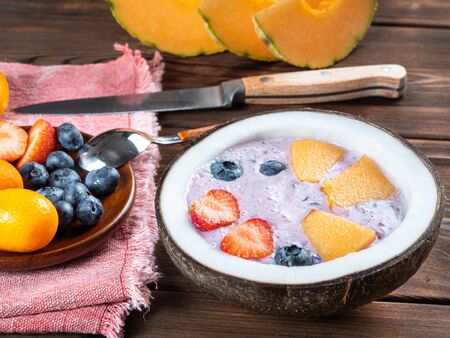 Close up view of blueberry smoothie in a coconut bowl with strawberries and melon and kumquat on a red towel with a teaspoon and knife.の写真素材