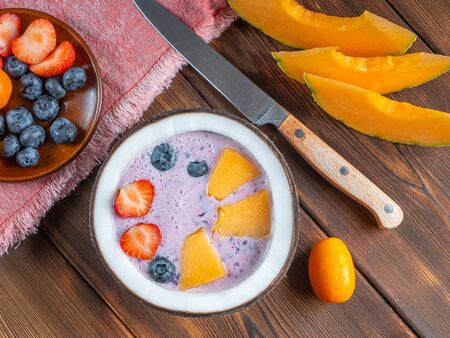 Blueberry smoothie in a coconut bowl with strawberries and melon and kumquat on a red towel with a teaspoon and knife.の写真素材