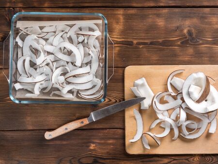 Top view of dried coconut chips in on baking paper in a glass pan and on cutting board with knifeの写真素材
