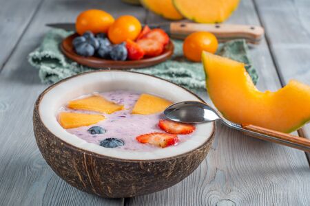 Close up view of blueberry smoothie in a coconut bowl with strawberries and melon and kumquat on a white wooden background with teaspoon and knife.. Soft focus.の写真素材