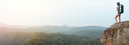 Young woman with backpack standing on cliff's edge and looking to a skyの写真素材