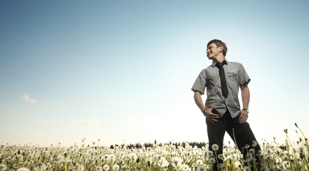 Young smiling man standing on a meadow with dandelions on a blue sky background