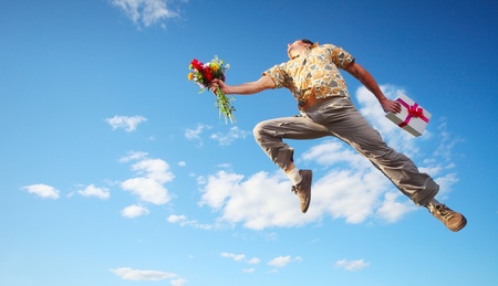 Young happy man jumping with flowers and gift box on blue sky backgroundの写真素材