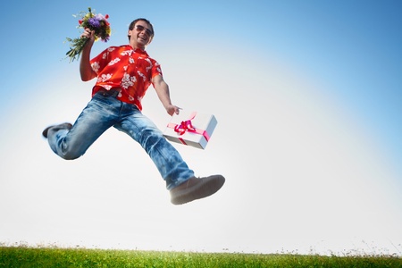 Young happy man in sunglasses jumping with flowers and gift box on blue sky backgroundの写真素材