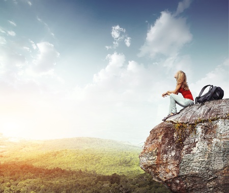 Young woman with backpack sitting on cliff's edge and looking to a sky with cloudsの写真素材