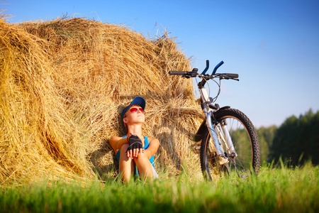 Young woman with bicycle sitting near straw heap and looking to a skyの写真素材